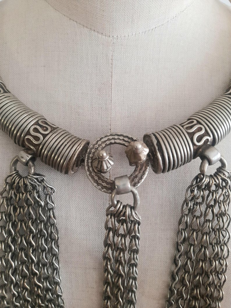 May include: Silver-toned necklace with intricate detailing. The necklace features a coiled design on the upper portion, connected to a central circular element. Dangling from the necklace are multiple chains, creating a layered effect. The piece is displayed on a mannequin.