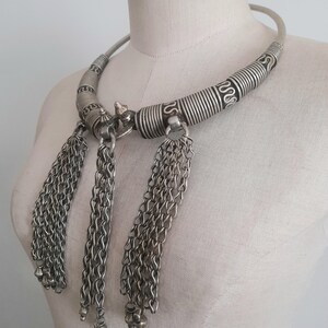 May include: A silver-toned necklace with a rigid collar and three chain tassel pendants. The collar features spiral and linear detailing. The tassels are composed of multiple chains ending in small, round beads. The necklace is displayed on a mannequin.