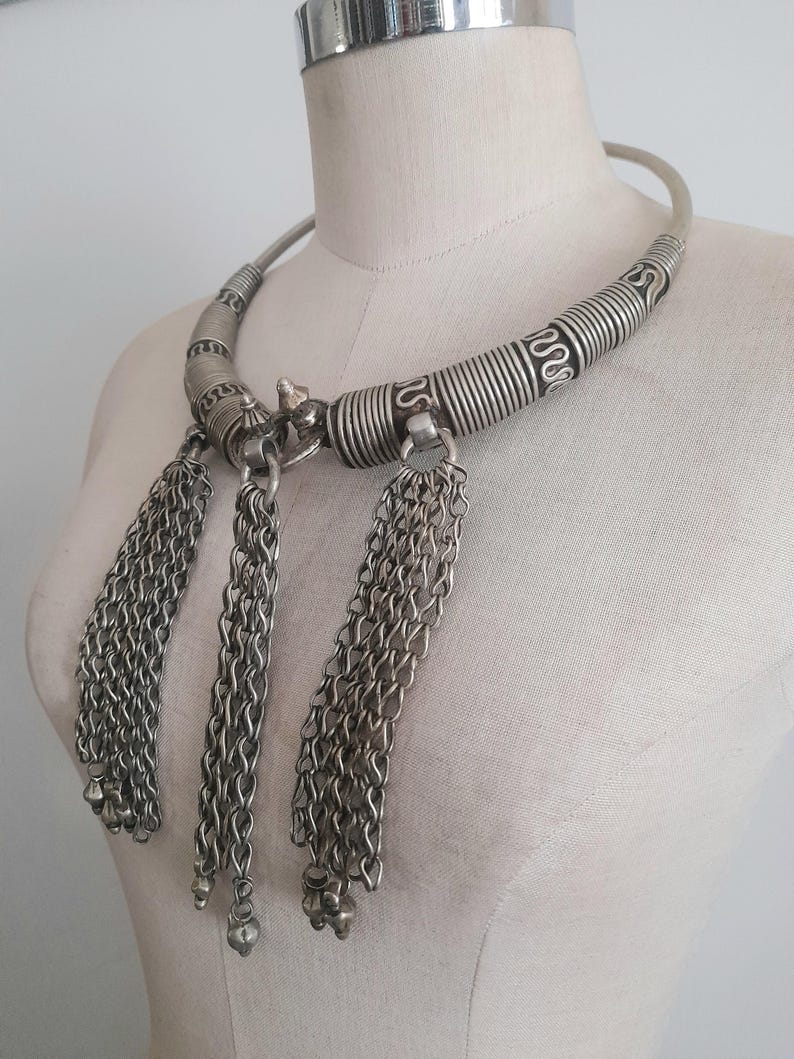 May include: A silver-toned necklace with a rigid collar and three chain tassel pendants. The collar features spiral and linear detailing. The tassels are composed of multiple chains ending in small, round beads. The necklace is displayed on a mannequin.