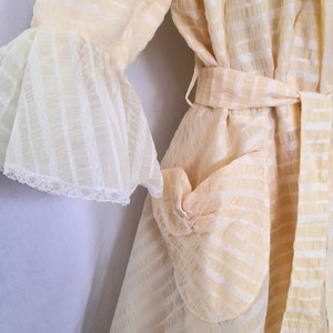 May include: A cream-colored robe with a white striped pattern and a matching belt. The robe has a ruffled sleeve and a patch pocket on the front.