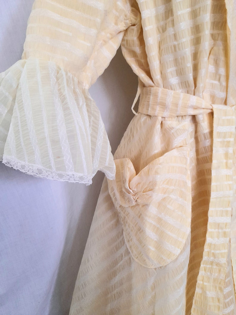 May include: A cream-colored robe with a white striped pattern and a matching belt. The robe has a ruffled sleeve and a patch pocket on the front.