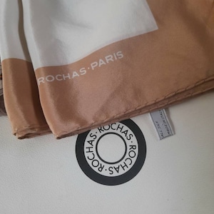 May include: A white silk scarf with a brown border and the text "ROCHAS PARIS" printed on it. The scarf is folded and the label is visible, which reads "ALL SILK MADE IN ITALY".