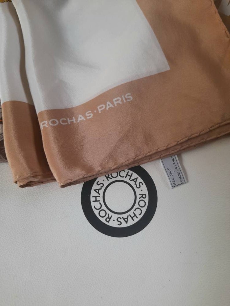 May include: A white silk scarf with a brown border and the text "ROCHAS PARIS" printed on it. The scarf is folded and the label is visible, which reads "ALL SILK MADE IN ITALY".