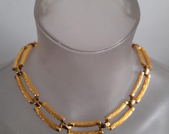 Puede incluir: Un collar dorado con un diseño de doble hebra. El collar presenta eslabones cilíndricos texturizados conectados por piezas rectangulares más pequeñas. El collar se exhibe sobre un busto transparente.