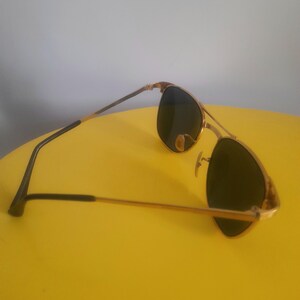 May include: Gold-framed sunglasses with dark lenses. The sunglasses have a classic aviator design with a double bridge and a gold finish on the frame. The temples are black and gold. The sunglasses are resting on a yellow surface.