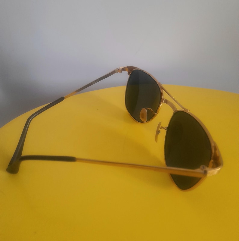 May include: Gold-framed sunglasses with dark lenses. The sunglasses have a classic aviator design with a double bridge and a gold finish on the frame. The temples are black and gold. The sunglasses are resting on a yellow surface.