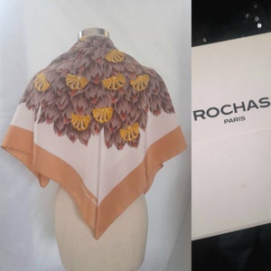 May include: A white silk scarf with a brown and yellow floral pattern. The scarf has a tan border and the text "ROCHAS PARIS" is printed on the border. The scarf is displayed on a white mannequin.