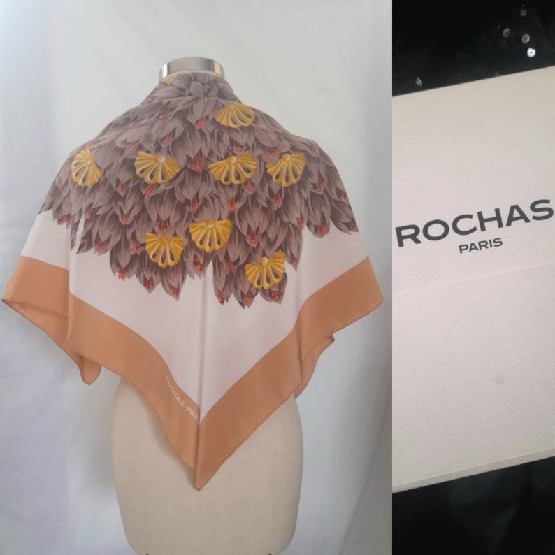 May include: A white silk scarf with a brown and yellow floral pattern. The scarf has a tan border and the text "ROCHAS PARIS" is printed on the border. The scarf is displayed on a white mannequin.