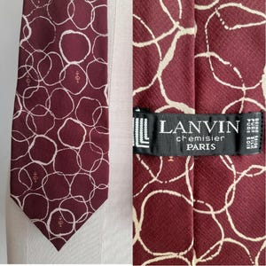 Könnte beinhalten: Eine burgunderfarbene Seidenkrawatte mit einem weißen abstrakten Muster. Die Krawatte hat ein Etikett mit der Aufschrift "LANVIN chemiseir PARIS" und "PURE SILK" in weißer Schrift auf schwarzem Hintergrund.