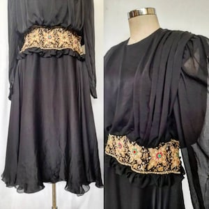 Puede incluir: Vestido negro con mangas largas y cinturón bordado. El cinturón presenta detalles dorados, rojos y verdes. El vestido tiene una falda fluida y un dobladillo con volantes. El vestido se exhibe en un maniquí.