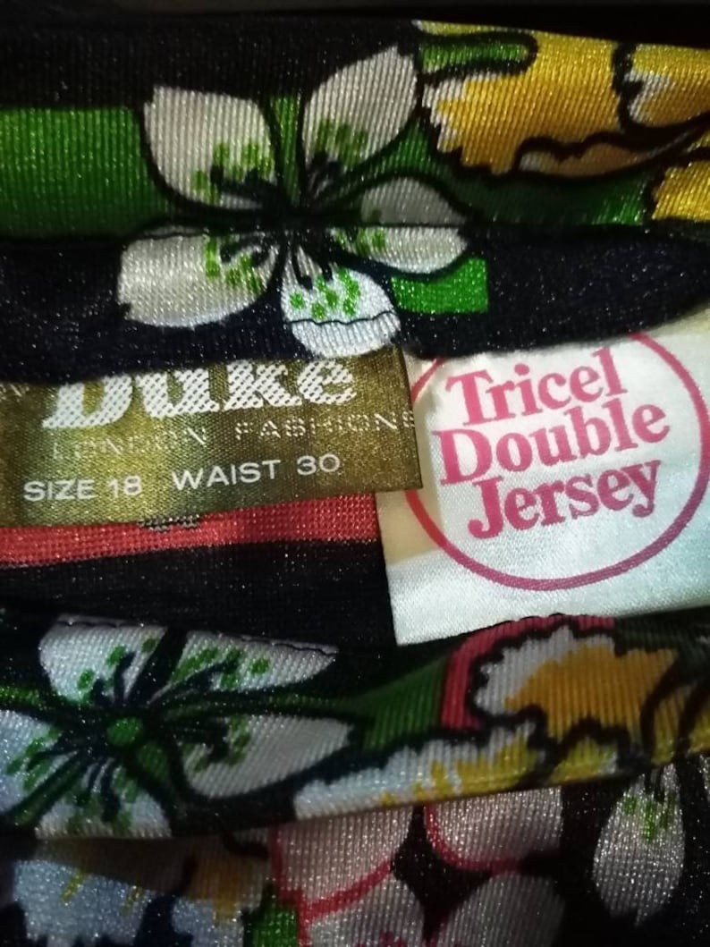 Pode incluir: Um tecido floral preto e branco com uma etiqueta dourada que diz "Duke London Fashions" e "Size 18 Waist 30". Uma etiqueta branca com letras cor-de-rosa diz "Tricol Double Jersey".