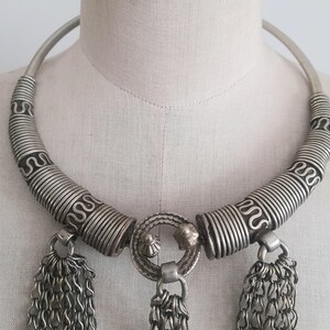 May include: Silver-toned statement necklace with a rigid collar and intricate detailing. The necklace features coiled wire accents and three chain tassel pendants. The central pendant has a circular design with small decorative elements.