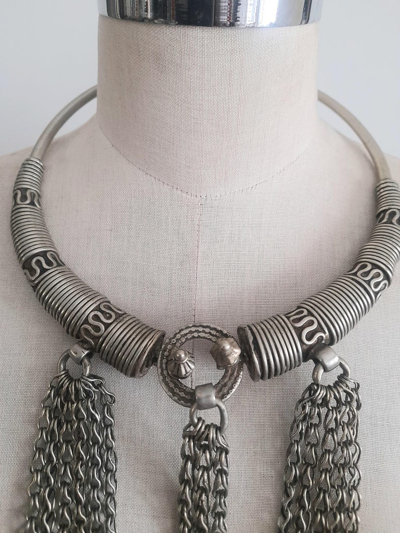 May include: Silver-toned statement necklace with a rigid collar and intricate detailing. The necklace features coiled wire accents and three chain tassel pendants. The central pendant has a circular design with small decorative elements.