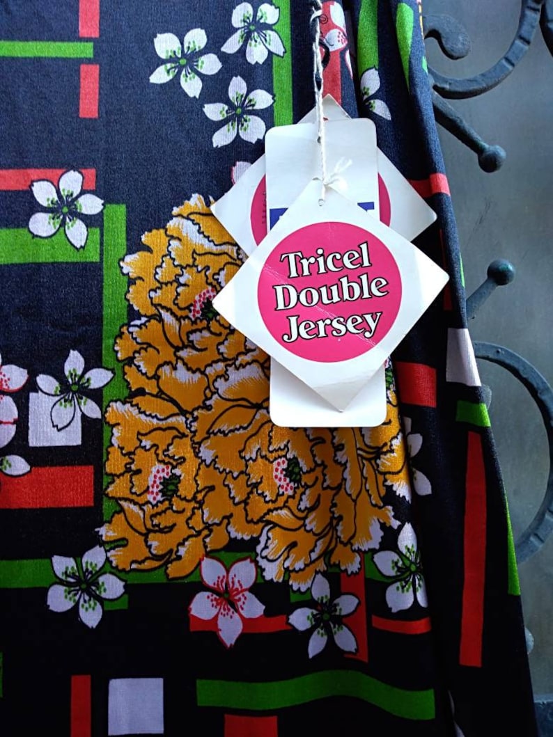 Pode incluir: Tecido preto com um padr&atilde;o floral em tons de vermelho, verde, branco e amarelo. O tecido tem uma etiqueta que diz "Tricel Double Jersey".