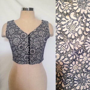 Puede incluir: Un top sin mangas, recortado, hecho de encaje blanco y negro con un patrón floral. El top tiene un escote en V y un cierre de botón en la parte delantera.