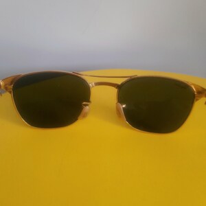 May include: Gold-framed sunglasses with dark lenses. The sunglasses have a double bridge and a classic aviator style. The lenses are a dark green color, and the frames have a shiny gold finish. The sunglasses are set against a yellow background.