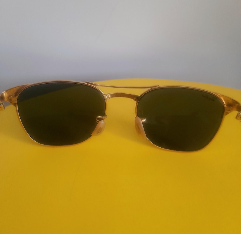 May include: Gold-framed sunglasses with dark lenses. The sunglasses have a double bridge and a classic aviator style. The lenses are a dark green color, and the frames have a shiny gold finish. The sunglasses are set against a yellow background.