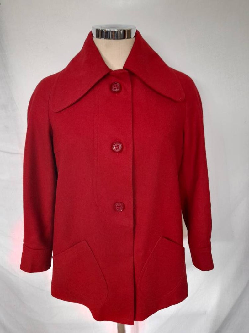 K&ouml;nnte beinhalten: Eine rote Wolljacke mit einem breiten Kragen und vier Kn&ouml;pfen auf der Vorderseite. Die Jacke hat zwei aufgesetzte Taschen auf der Vorderseite.