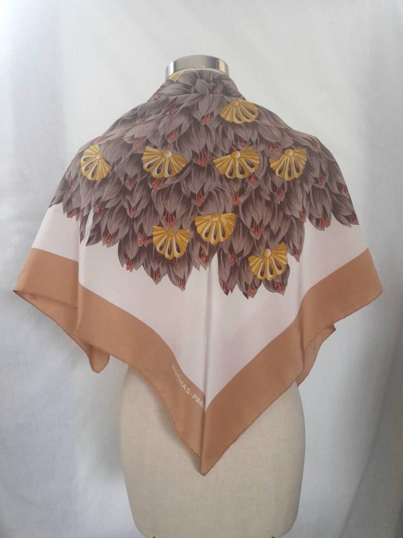 May include: A white silk scarf with a brown and yellow floral design. The scarf has a brown border and the text "ROCHAS - PARIS" is printed in the lower right corner.