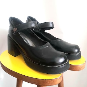May include: A pair of black Mary Jane shoes featuring a thick platform sole, a chunky block heel, and a single adjustable strap across the instep secured with a small metallic button detail. The shoes have a smooth finish and a rounded toe design.