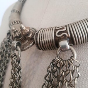 May include: Close-up of a silver-toned necklace with intricate details. The necklace features a thick, coiled collar and multiple strands of chain. Decorative elements include a small, ornate clasp and a textured design.