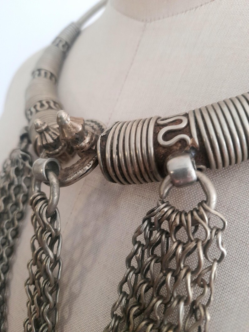 May include: Close-up of a silver-toned necklace with intricate details. The necklace features a thick, coiled collar and multiple strands of chain. Decorative elements include a small, ornate clasp and a textured design.