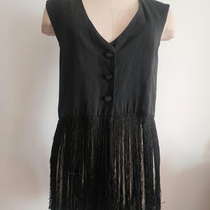 Peut inclure: Robe noire sans manches avec un décolleté en V et trois boutons sur le devant. La robe présente des franges au niveau de l'ourlet. Le vêtement est présenté sur un mannequin.