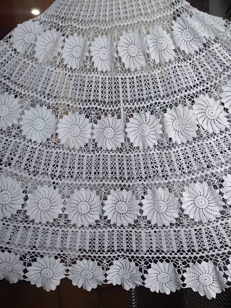 May include: White crocheted tablecloth with a delicate floral pattern. The tablecloth features a repeating design of small, intricate flowers.