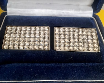 May include: Two rectangular silver cufflinks, each encrusted with numerous clear, sparkling crystals. The cufflinks are displayed inside a dark blue velvet box with a white satin lining, suggesting a presentation or storage case.