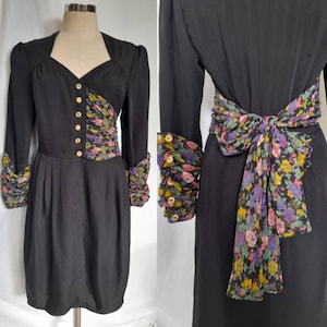May include: Black vintage dress with long sleeves, a V-neck, and a pleated skirt. The dress features a floral print on the bodice, cuffs, and a bow at the waist. Gold buttons adorn the front of the dress, adding a touch of elegance to the design.