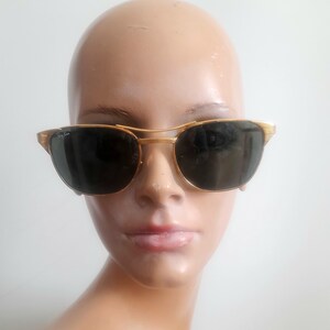 May include: Gold-framed sunglasses with dark lenses. The sunglasses have a double bridge and a classic design. The frame is a shiny gold color, and the lenses are a dark, solid color. The sunglasses are on a mannequin head.