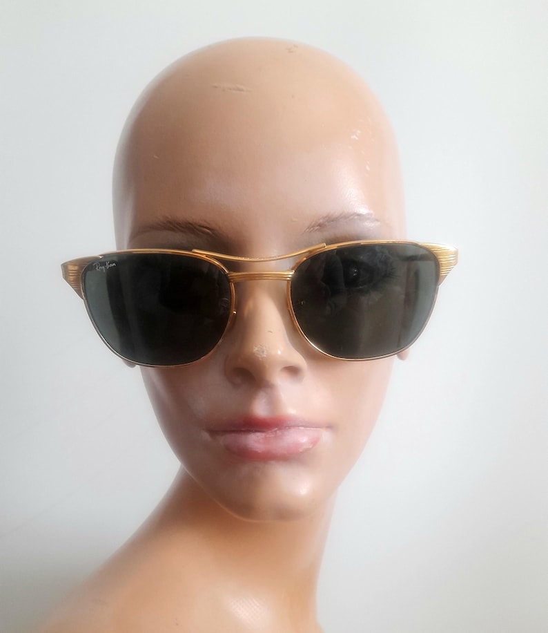 May include: Gold-framed sunglasses with dark lenses. The sunglasses have a double bridge and a classic design. The frame is a shiny gold color, and the lenses are a dark, solid color. The sunglasses are on a mannequin head.