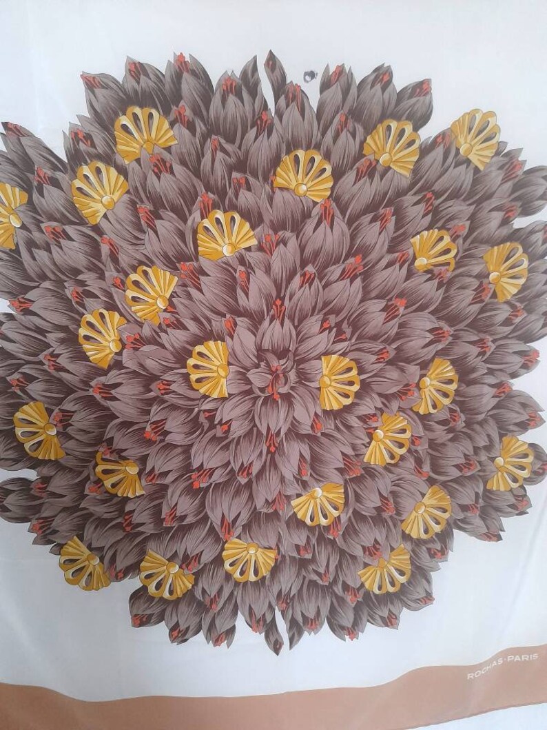 May include: A white silk scarf with a brown and yellow floral design. The design features a large, circular pattern of stylized flowers with yellow centers and brown petals. The scarf has a brown border with the text "ROCHAS PARIS" in white.
