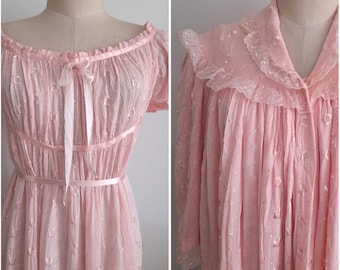 Op de afbeelding: Een vintage roze nachtjapon en badjas set. De nachtjapon heeft een strik, een getailleerde taille en een gerimpelde halslijn. De badjas heeft een met kant afgezette kraag en een knoopsluiting. Beide items zijn gemaakt van een doorschijnende, gestippelde stof.