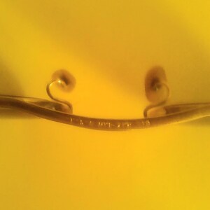 May include: A gold-colored, curved metal object with two decorative swirls at each end. The object has a series of small markings along its length. The background is a solid yellow color.