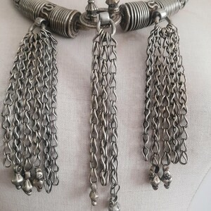 May include: Silver-toned necklace with a rigid, curved collar and three dangling chain tassels. The collar features coiled and patterned sections. Each tassel ends with small, decorative charms. The necklace is displayed on a mannequin.