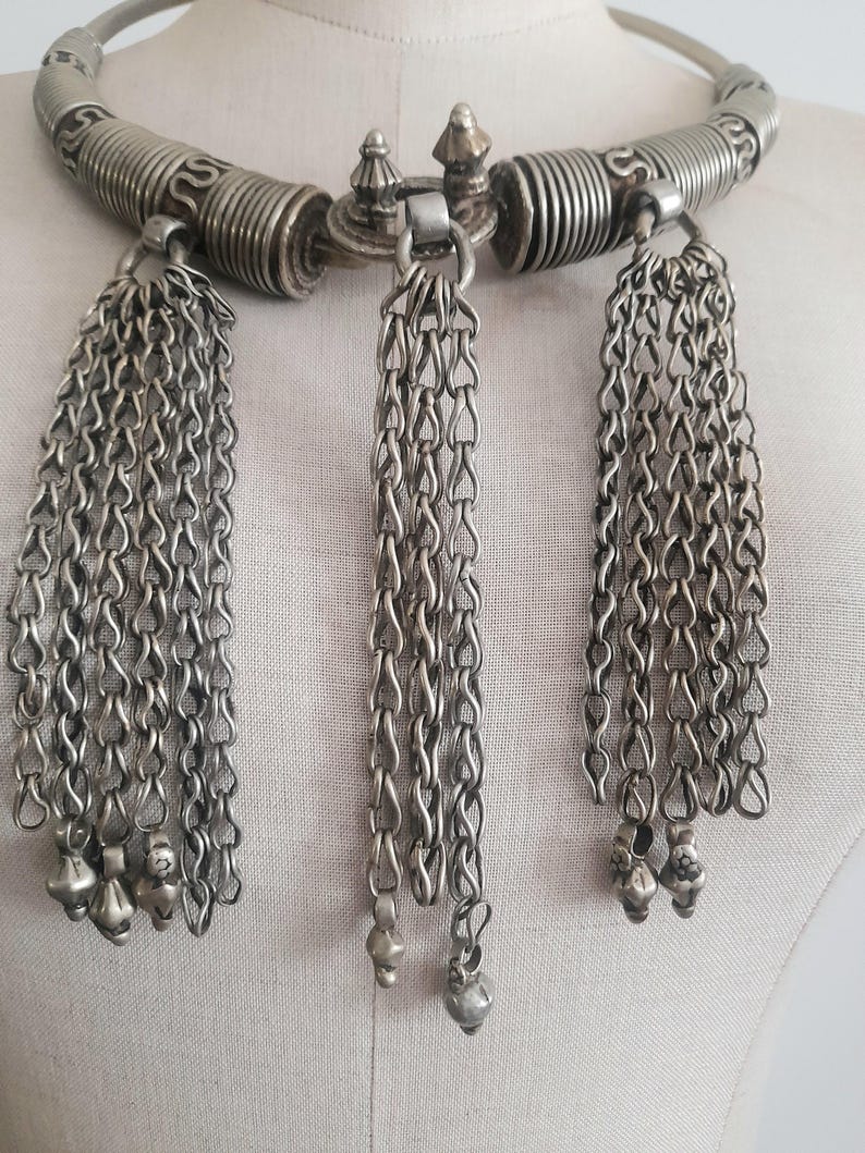 May include: Silver-toned necklace with a rigid, curved collar and three dangling chain tassels. The collar features coiled and patterned sections. Each tassel ends with small, decorative charms. The necklace is displayed on a mannequin.
