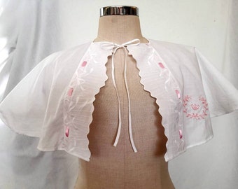 May include: White cotton cape with pink embroidered details and a scalloped edge. The cape has a tie at the neck.