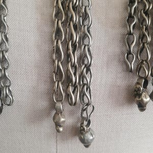 May include: Close-up of silver-toned metal chains with a series of linked loops. The chains have small, decorative pendants at the ends. The background is a light, neutral fabric.