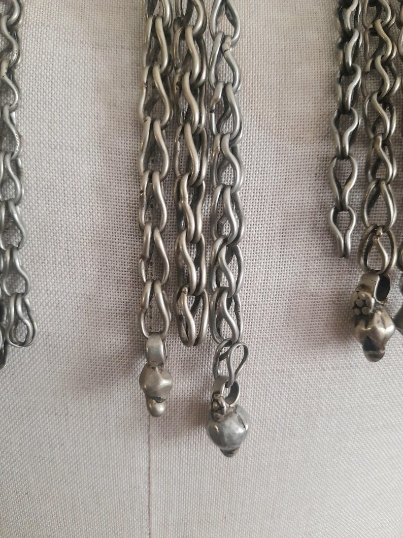 May include: Close-up of silver-toned metal chains with a series of linked loops. The chains have small, decorative pendants at the ends. The background is a light, neutral fabric.