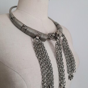 May include: Silver-toned statement necklace with a wide, textured collar and three chain tassel pendants. The necklace features intricate detailing and a vintage aesthetic, displayed on a mannequin.