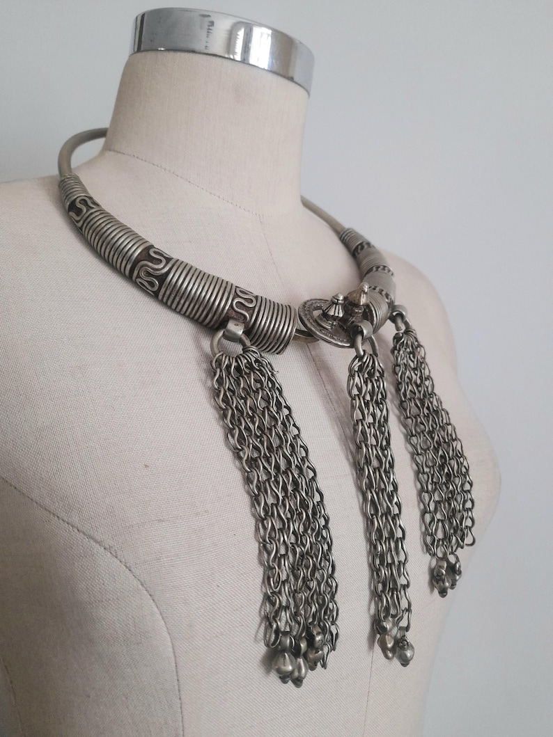 May include: Silver-toned statement necklace with a wide, textured collar and three chain tassel pendants. The necklace features intricate detailing and a vintage aesthetic, displayed on a mannequin.