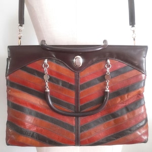 May include: A rectangular leather handbag featuring a chevron pattern of alternating dark brown, orange, and red stripes. The bag includes a dark brown top handle, a detachable shoulder strap, silver-toned hardware, and a central oval metal emblem.