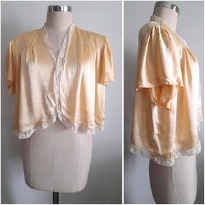 Può includere: Bolero vintage in raso color pesca con maniche corte. La giacca presenta un delicato bordo in pizzo bianco lungo i bordi e lo scollo. Il tessuto ha una sottile lucentezza e il capo è esposto su un manichino.