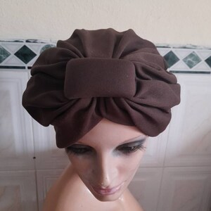 May include: A brown turban style hat with a large bow on top. The hat is made of a soft, stretchy fabric.