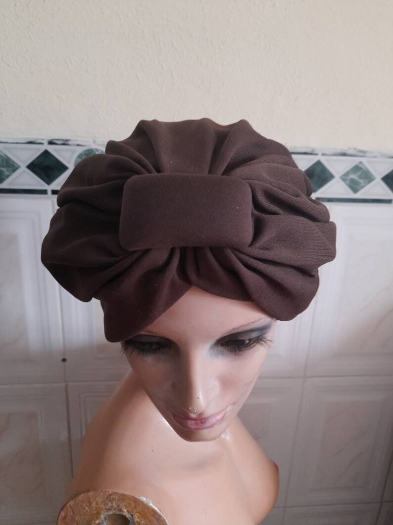 May include: A brown turban style hat with a large bow on top. The hat is made of a soft, stretchy fabric.