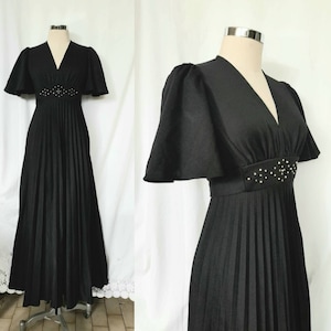 Könnte beinhalten: Schwarzes, bodenlanges Kleid mit V-Ausschnitt und plissiertem Rock. Das Kleid hat kurze, ausgestellte Ärmel und eine Perlenverzierung an der Taille. Das Kleid wird auf einer Schaufensterpuppe präsentiert.