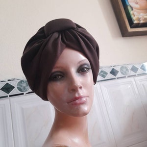 May include: A brown turban style head wrap made of fabric. The fabric is gathered and knotted at the top of the head.