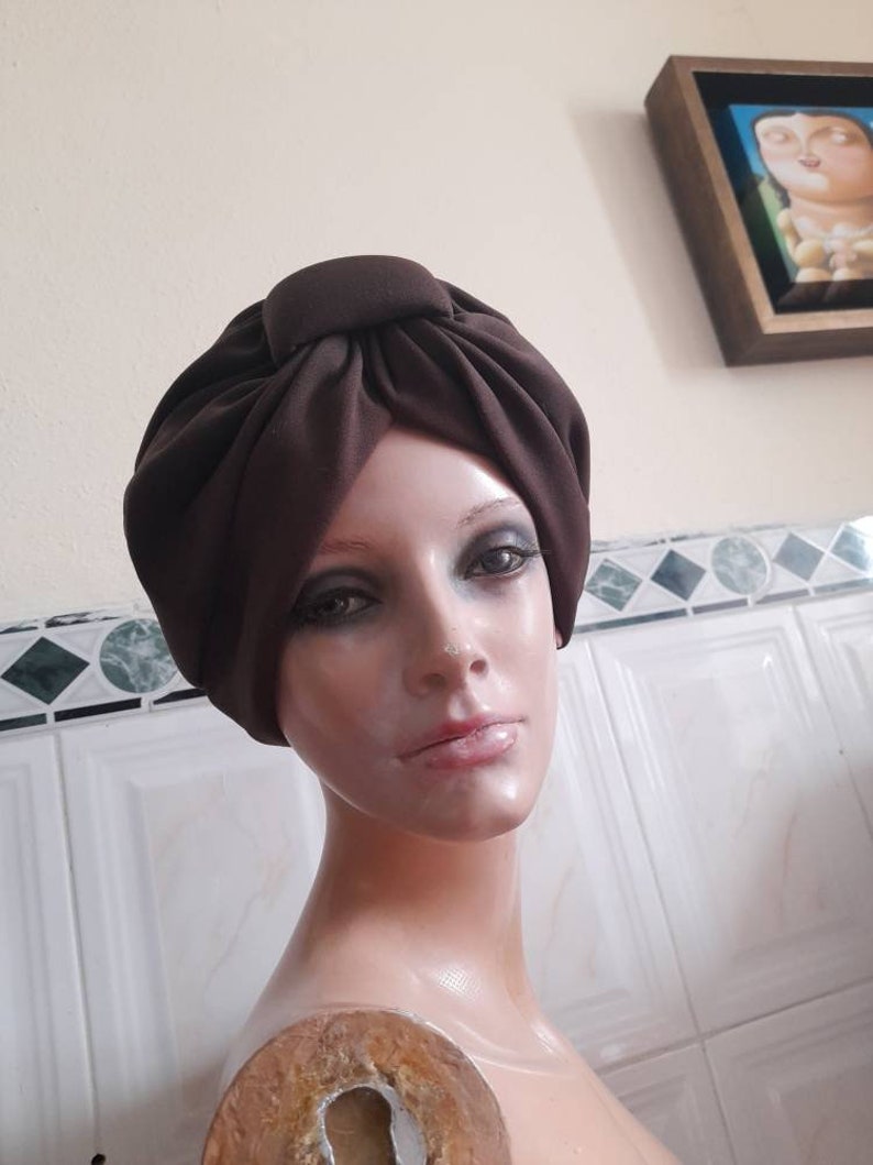 May include: A brown turban style head wrap made of fabric. The fabric is gathered and knotted at the top of the head.