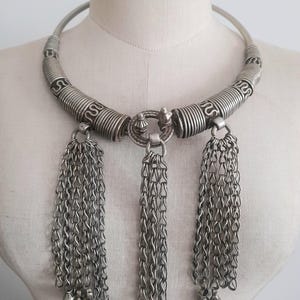 May include: Silver-toned statement necklace with a rigid collar and three chain tassel pendants. The collar features spiral and linear detailing. The tassels are composed of delicate chain links and small bell-shaped charms.